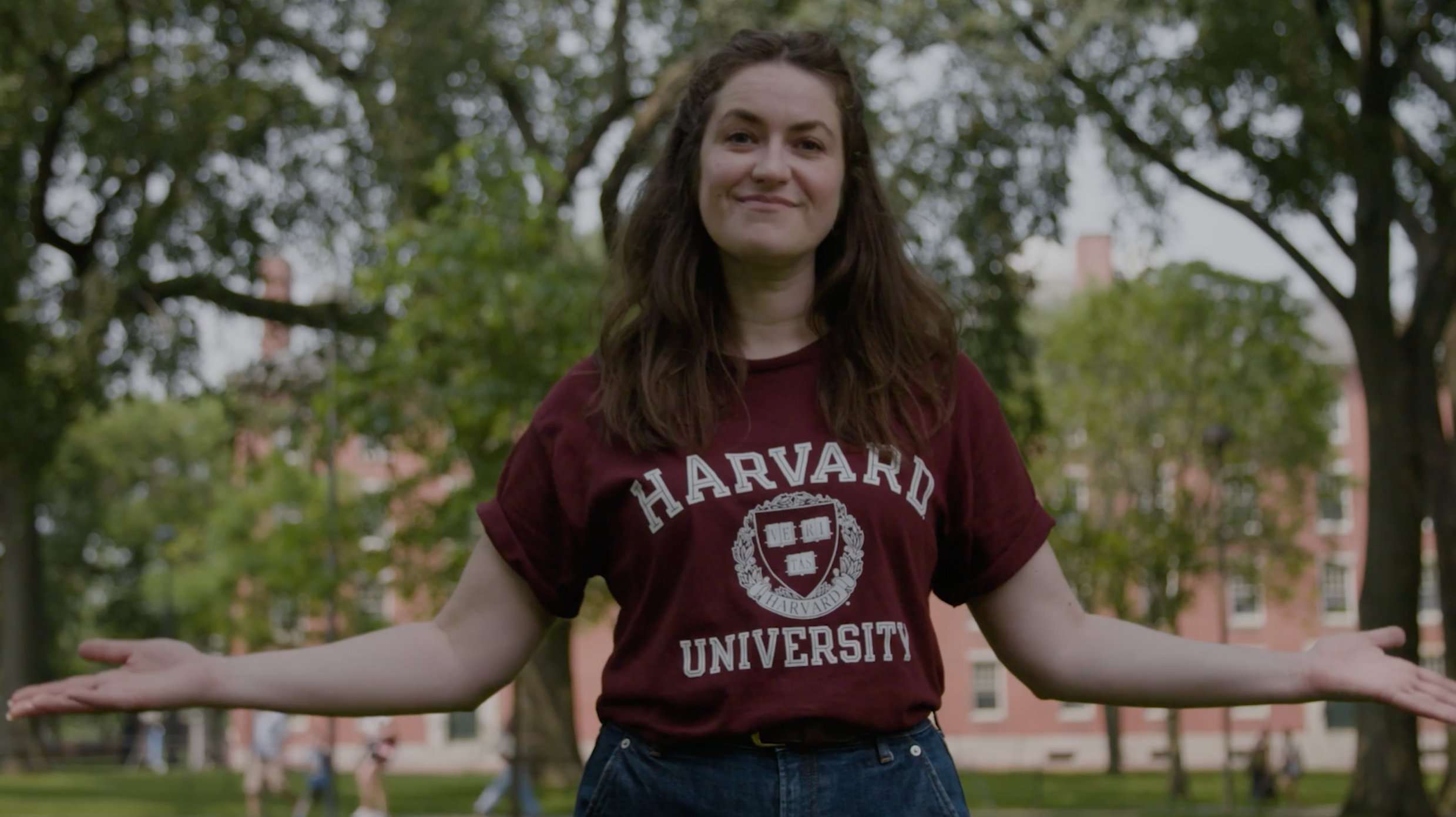 A student wearing a Harvard University tee shirt standing with her arms out on campus.