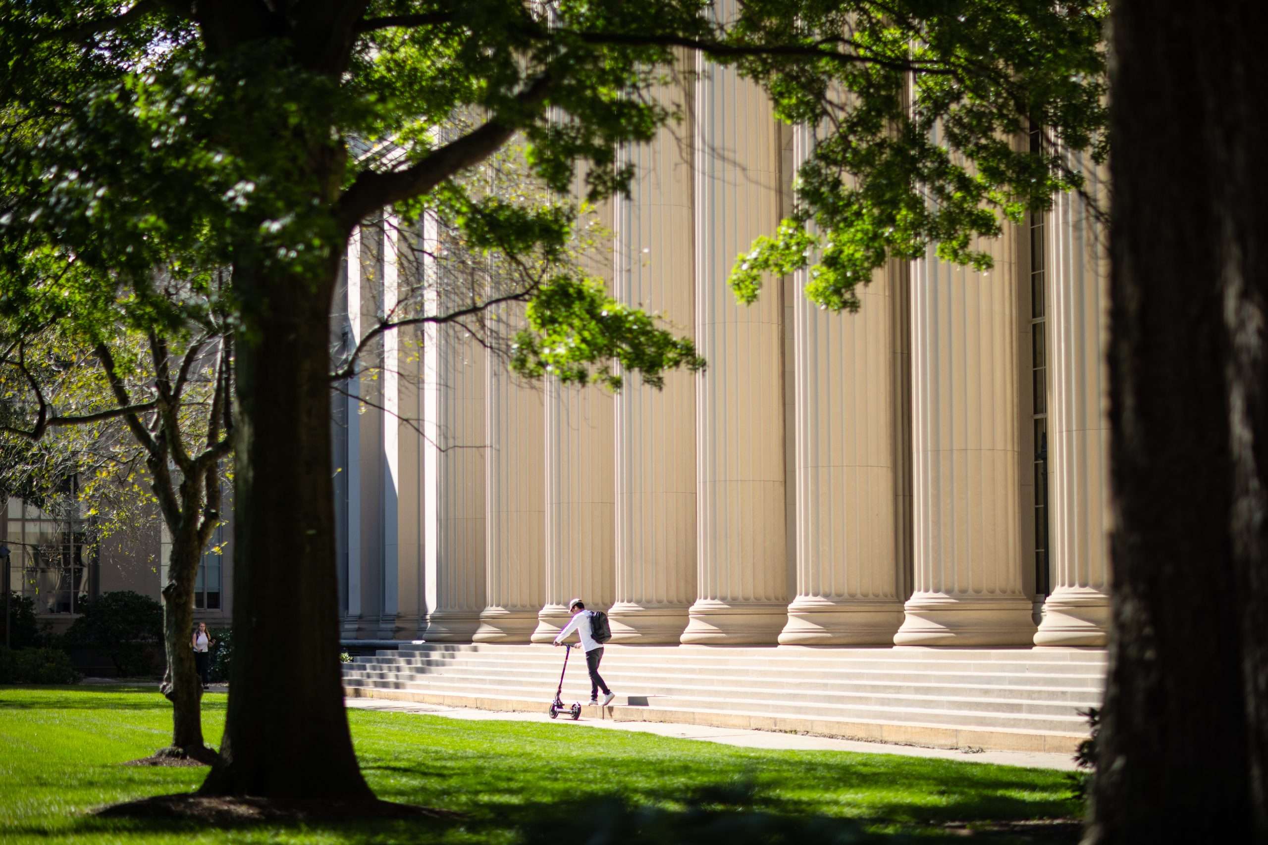 A person rides a scooter in front of the white steps of a columned building at the Massachusetts Institute of Technology.