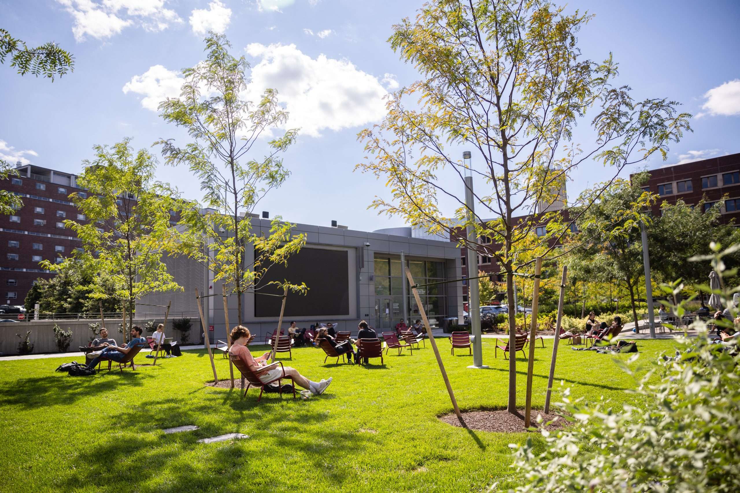 Students recline in red chairs on a campus lawn in front of a Brutalist building on a sunny day.